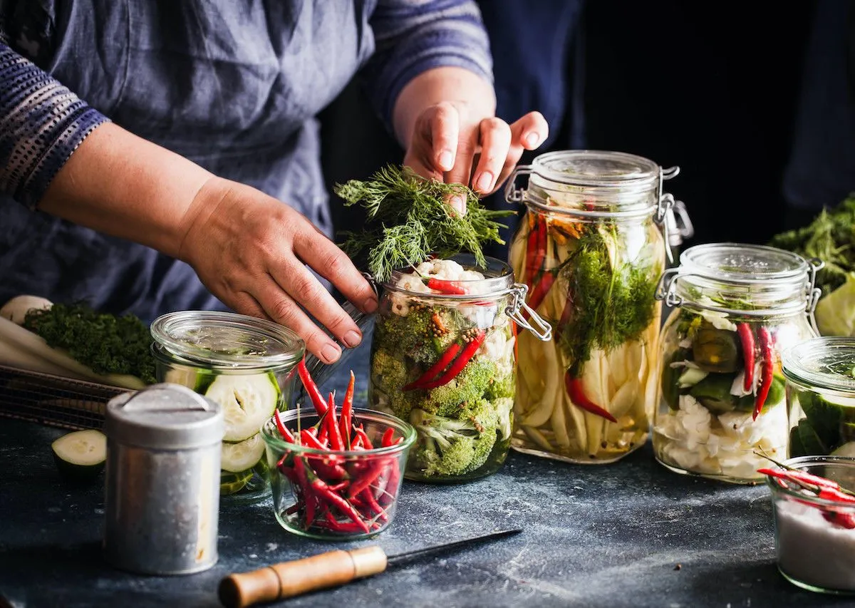 Hands putting food into jars surrounds by other jars with pickling vegetables in them. 
